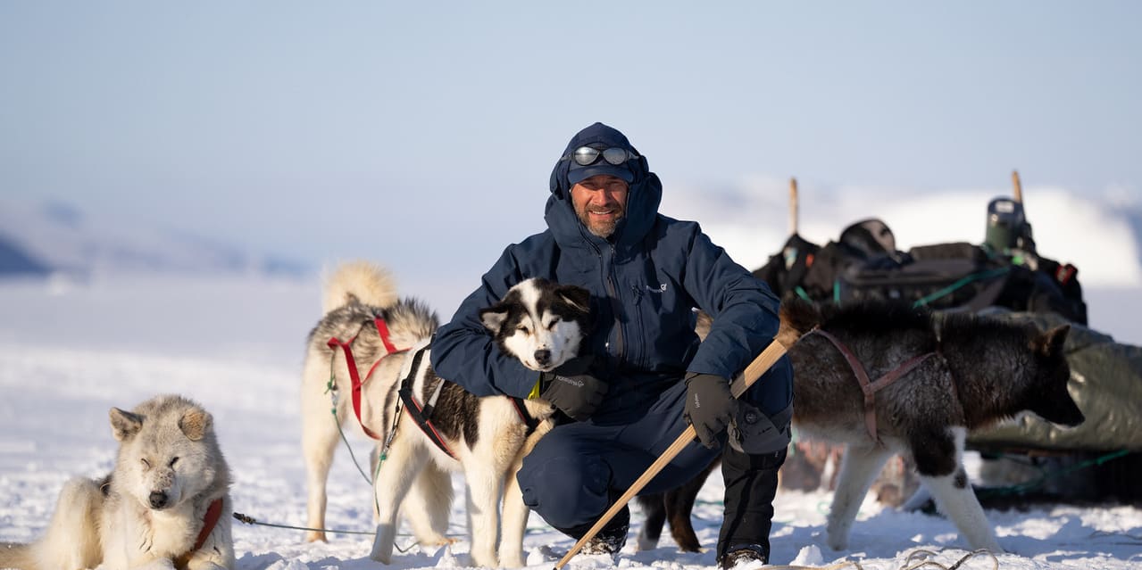 A man in a Norrøna jacket is sitting in the snow with a black and white dog.  Three other sled dogs are in the background. 
