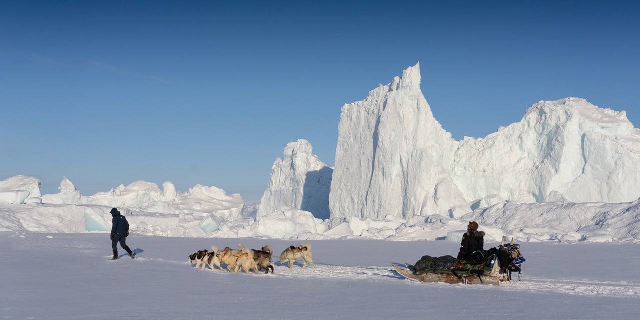 A person walks on a snowy landscape, a dog sledding team with a Norrøna backpack follows. The team is in front of a large ice berg. 
