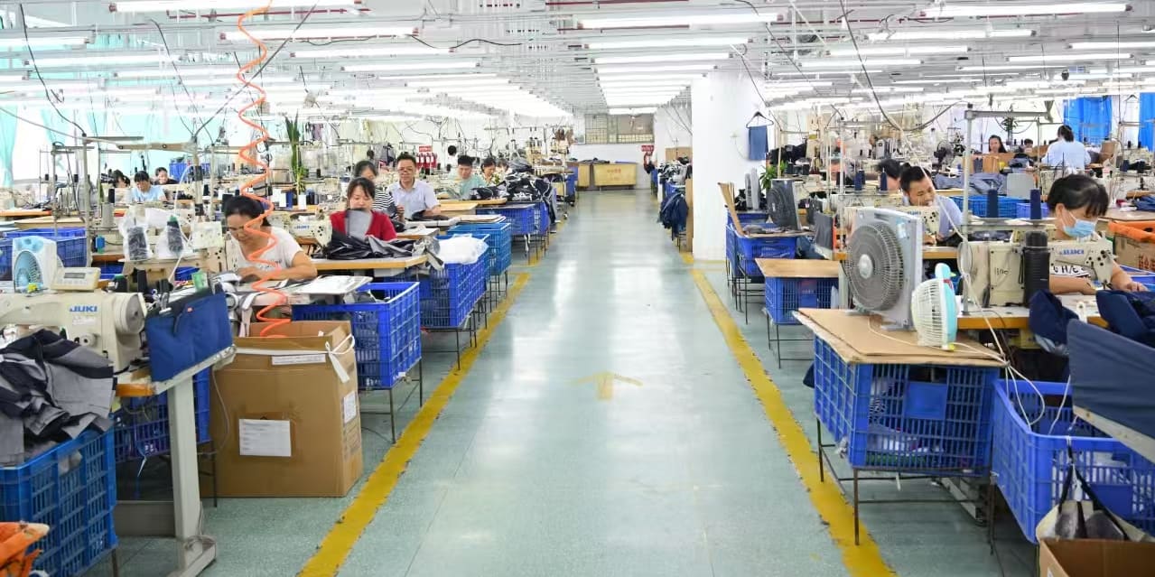 A brightly lit clothing factory with rows of workers at sewing machines, producing Norrøna apparel. Blue bins are present throughout the production line.
