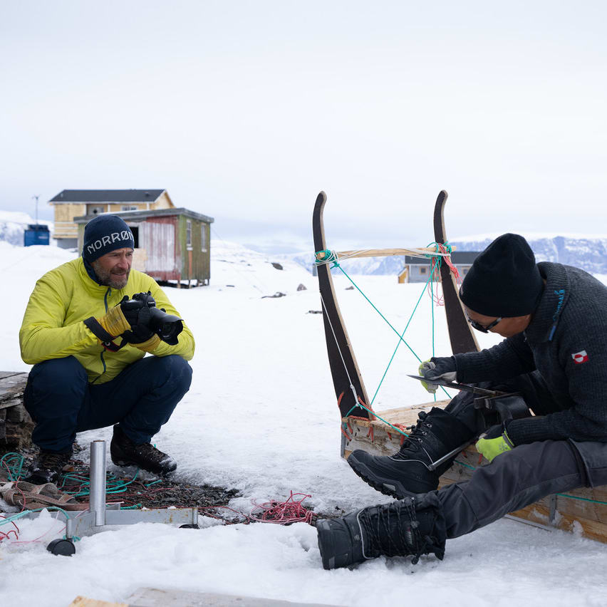 Two men, one wearing a Norrøna jacket, work on a sled in a snowy landscape. 
