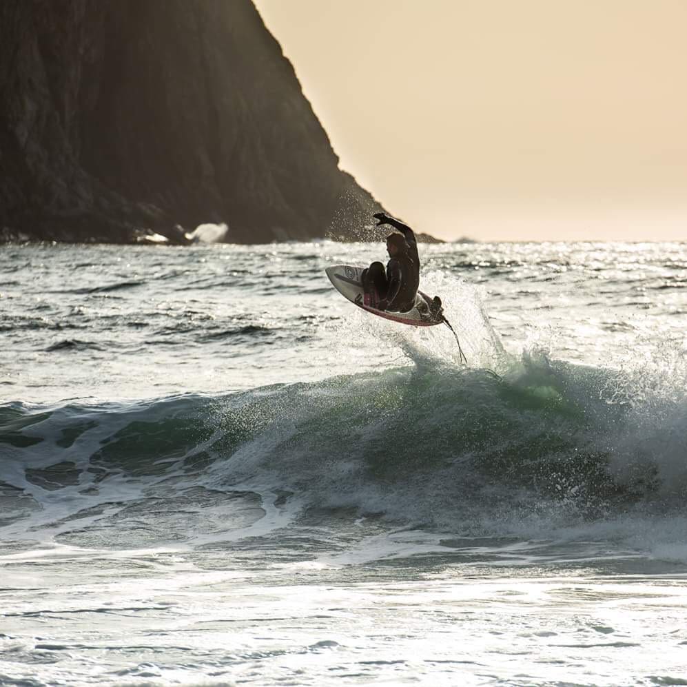 A surfer in a Norrøna wetsuit launches off a wave. The sun is setting behind the surfer, silhouetting a rocky headland in the background.