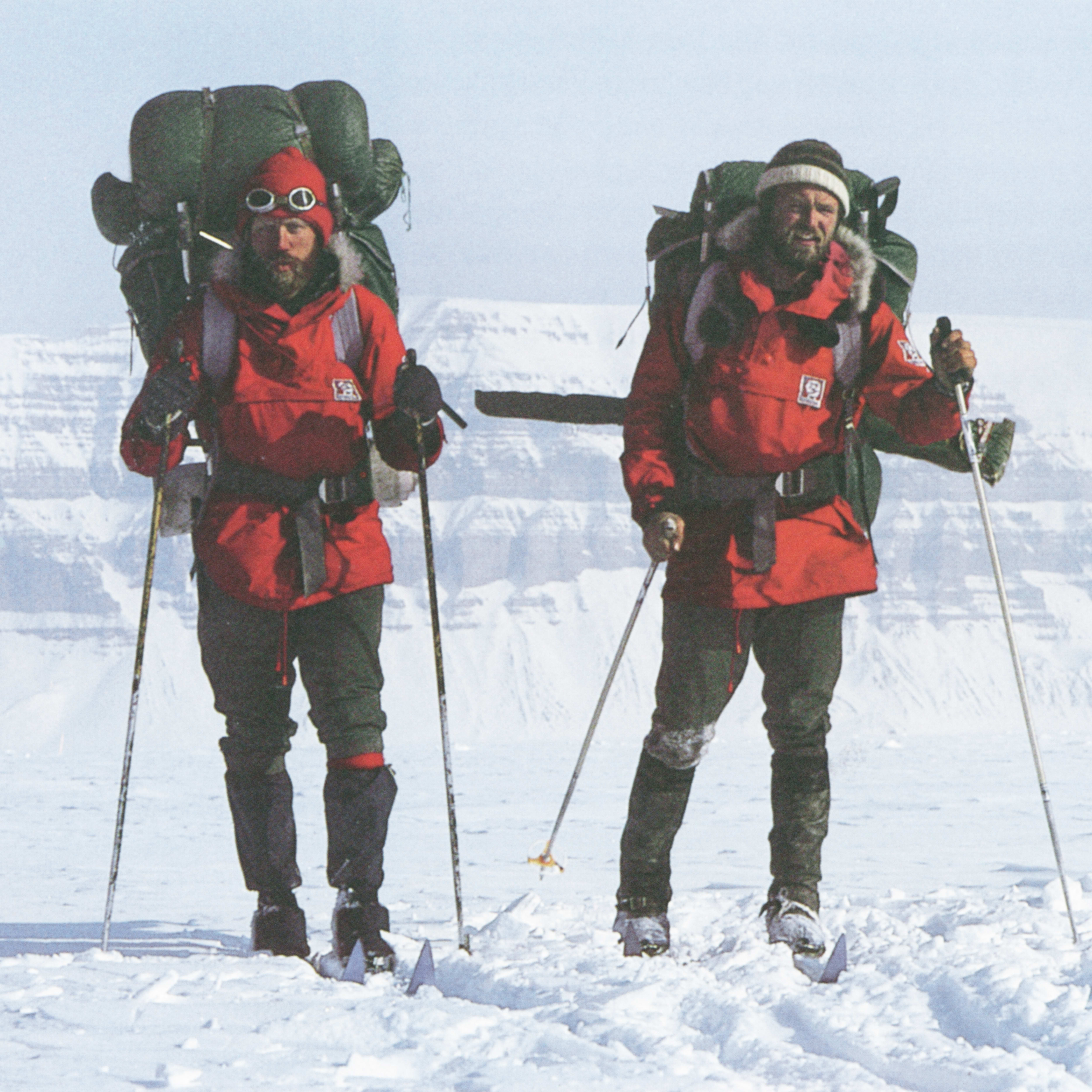 Two men are cross-country skiing in a snowy, mountainous landscape. Both men are wearing red jackets, backpacks, and ski poles. The sky is a light blue.