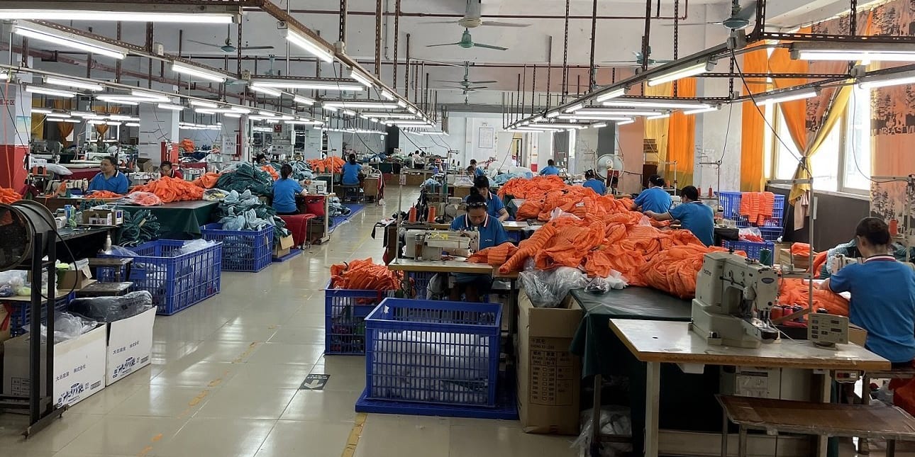 A large factory room with many people working at sewing machines. They are all wearing blue shirts and working on orange fabric. There are many blue plastic crates filled with fabric around the room.