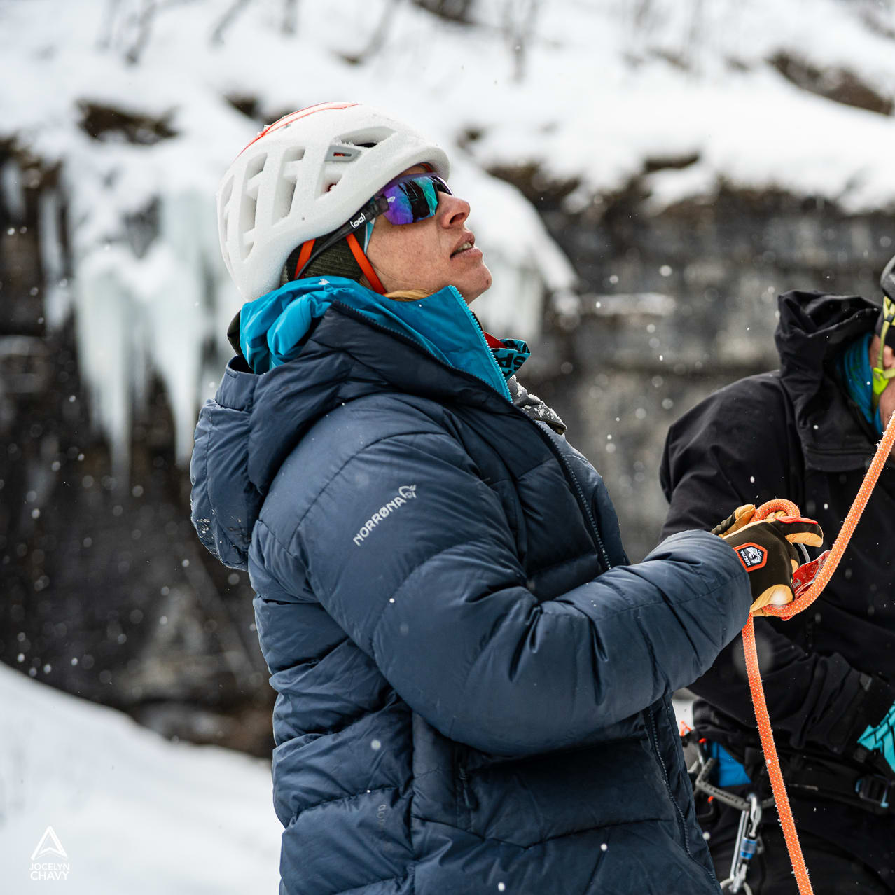 Two ice climbers in helmets use a rope on a snowy cliff. One wears a Norrøna puffer jacket and sunglasses.
