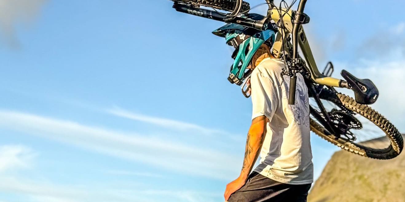 A person with a mountain bike over their shoulder wearing Norrøna pants and a helmet stands on a grassy hillside overlooking a valley.