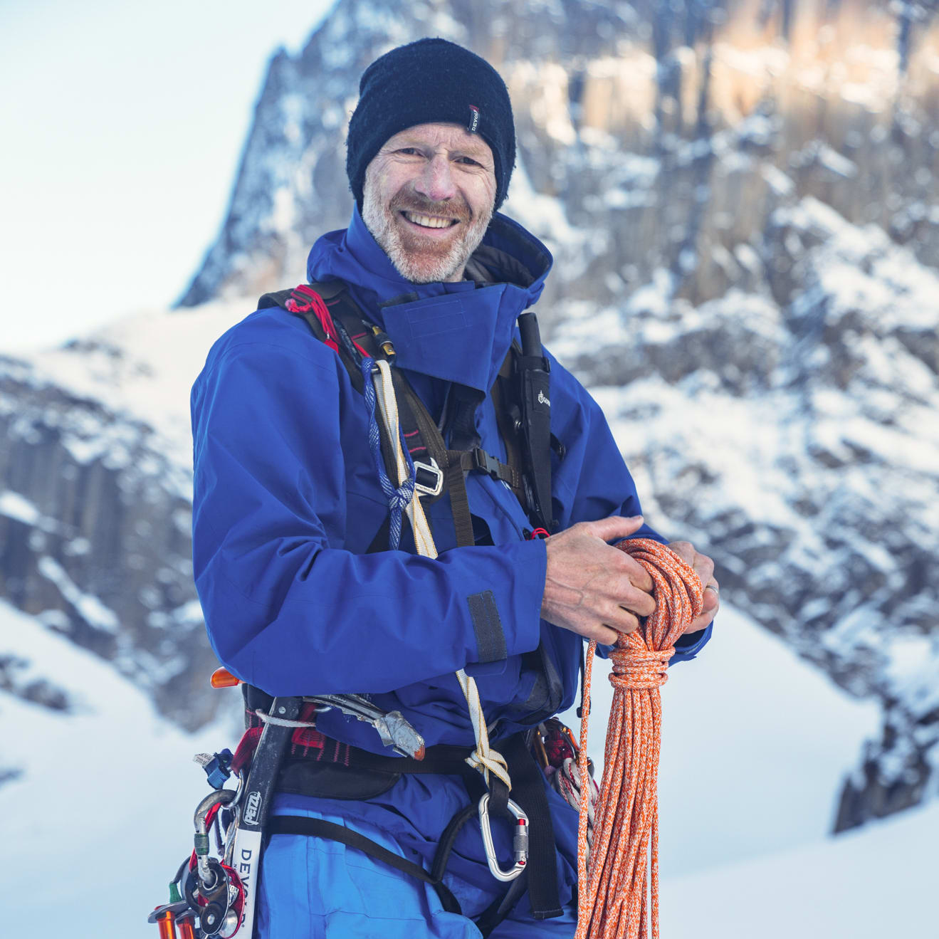 A smiling climber in Norrøna blue outerwear, harness and a black beanie, holds an orange climbing rope against a snowy mountain backdrop.
