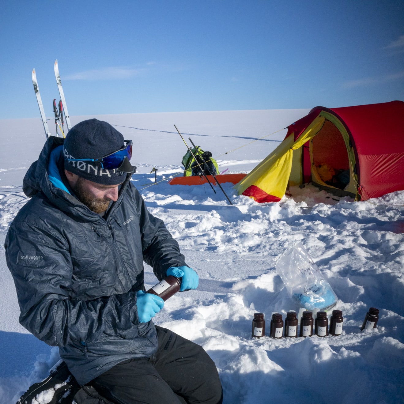 A person kneeling in snow holding a bottle while wearing a Norrøna jacket and gloves. A tent and skis are visible in the background.