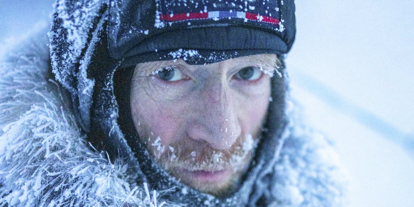 Close-up of a man wearing a fur-lined Norrøna jacket and hat covered in frost. He has light eyes, a beard, and is staring intensely at the camera.