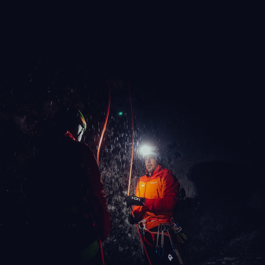 Two climbers in Norrøna gear, illuminated by headlamps, stand on an ice wall. 
