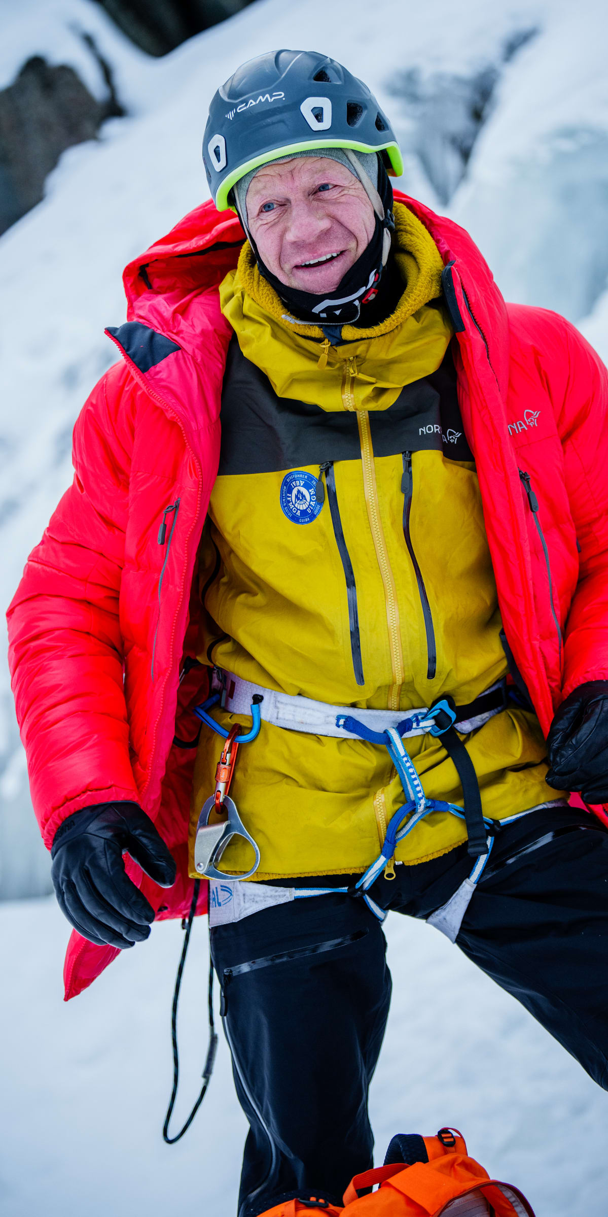 A man wearing a Norrøna jacket and pants, a helmet, and gloves stands in the snow. He is smiling.