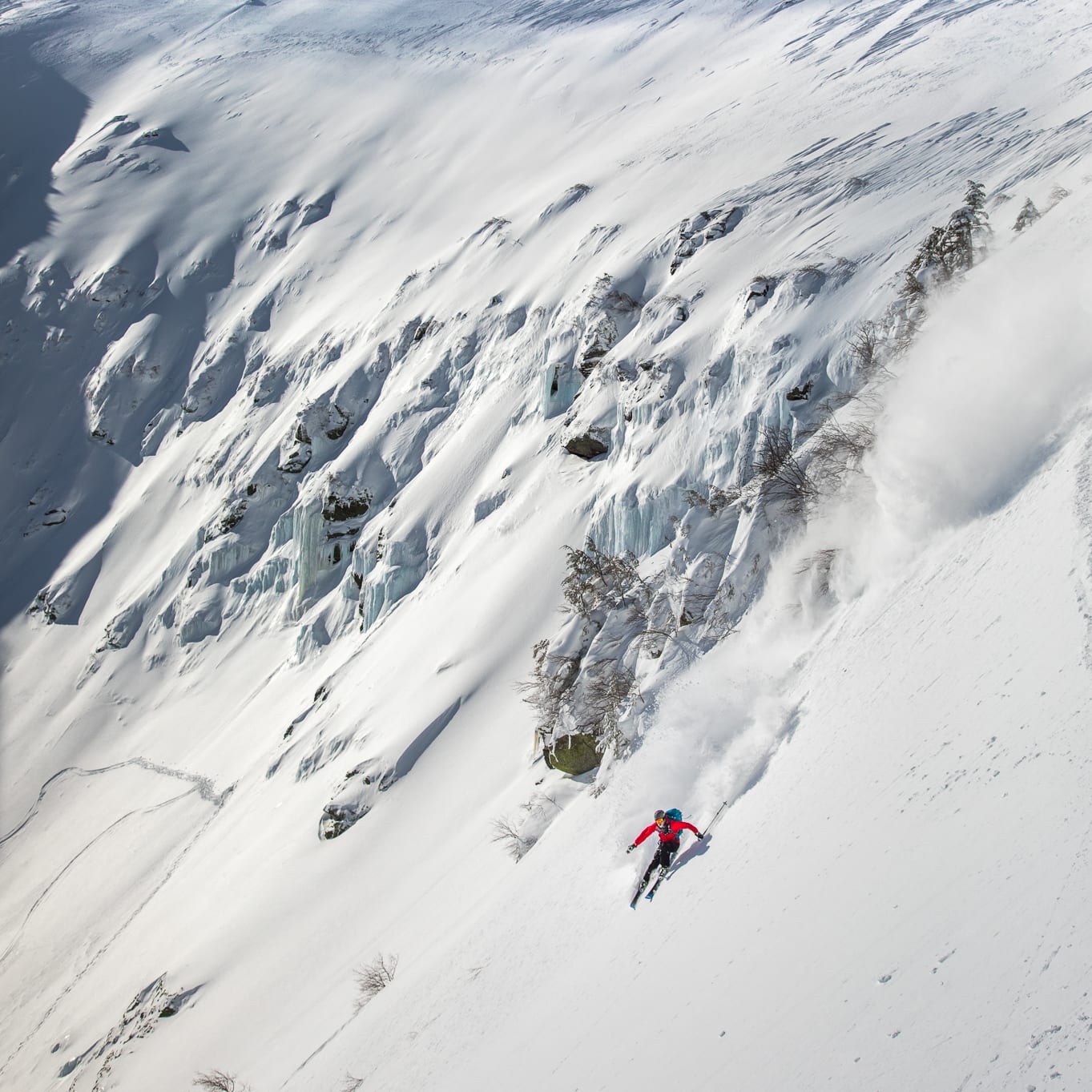 A skier in red Norrøna gear makes a turn down a snowy mountain slope.  The skier is leaving a trail of snow behind them.