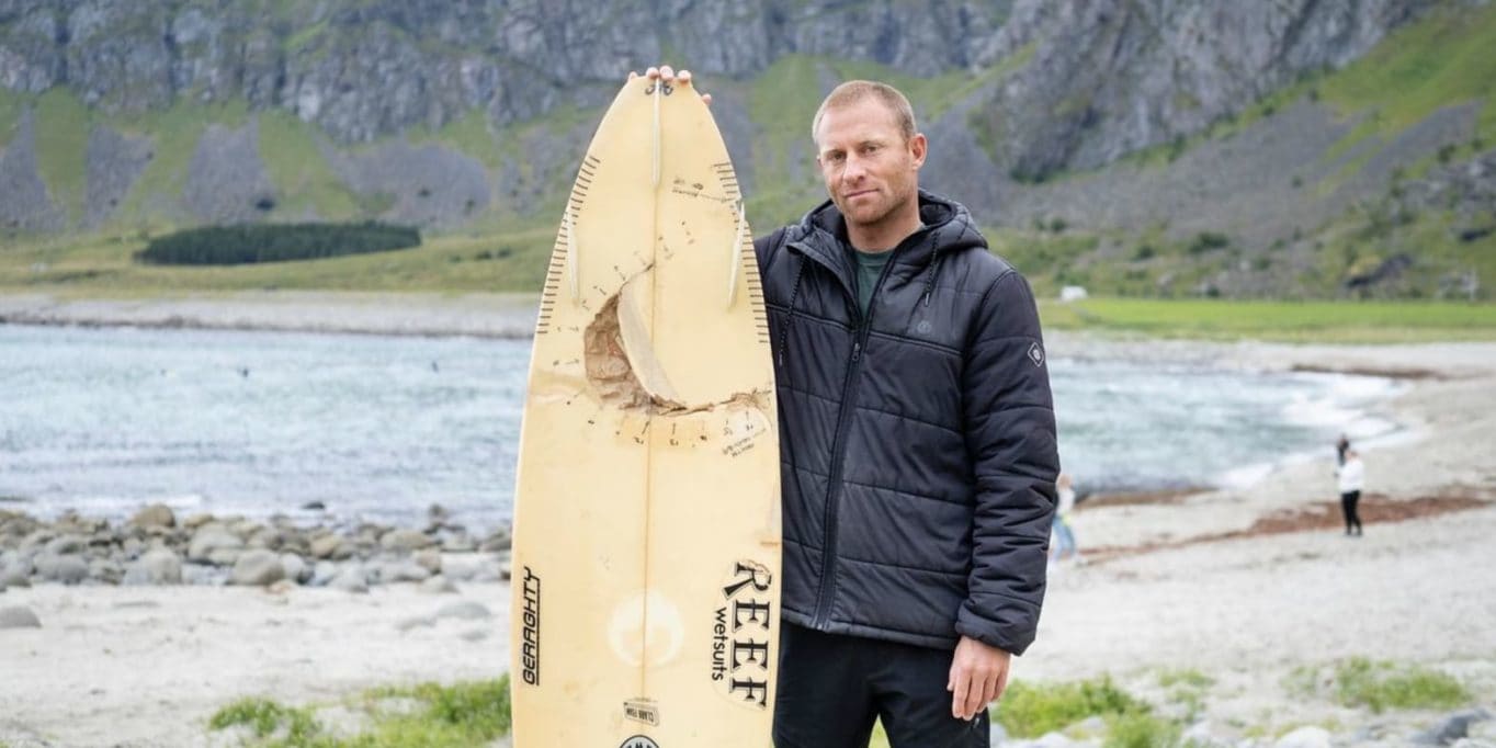 Man standing on a beach holding a surfboard. He is wearing a black Norrøna jacket. The background is a mountain range.