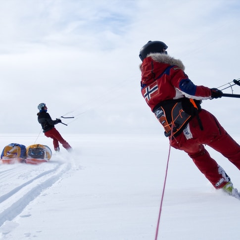 Two snowkiters on a snowy plain. The person in the foreground wears red Norrøna outerwear and skis, pulling sleds.