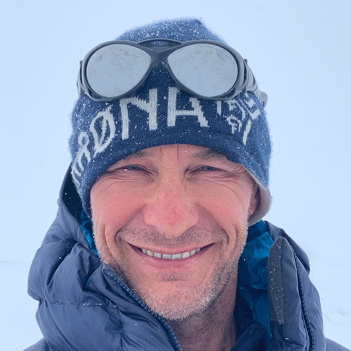 Close-up of a smiling man outdoors in snow, wearing a Norrøna hat and a puffy jacket. Goggles are pushed up on the hat.