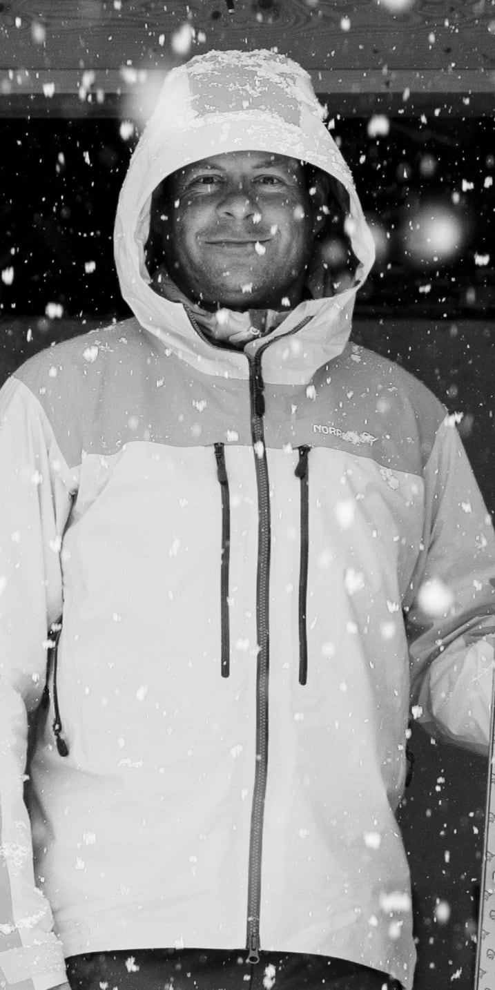 A man in a Norrøna jacket stands in the snow, holding a ski. 
