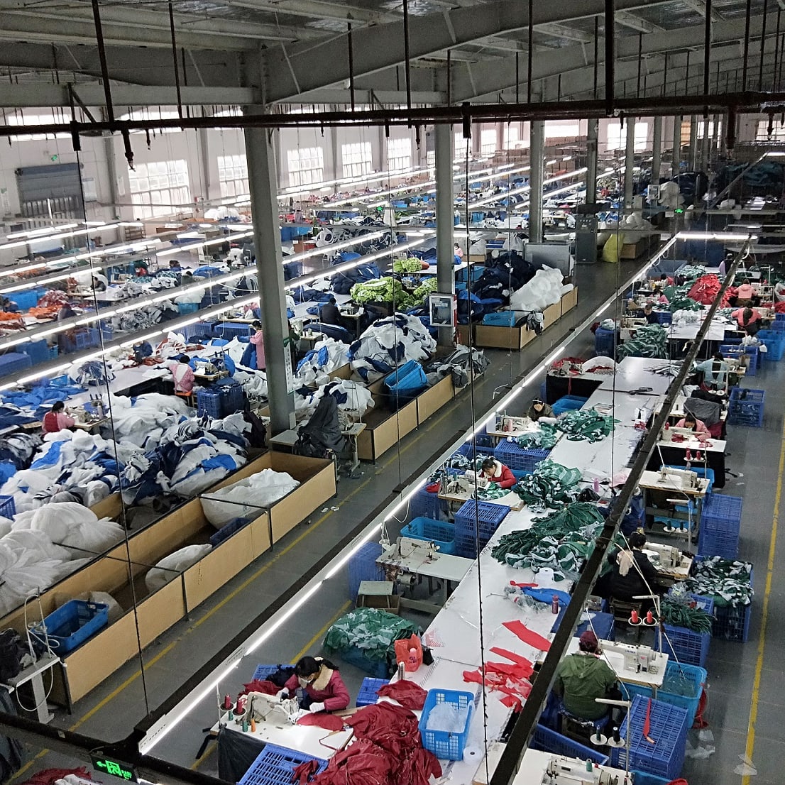 A large factory workspace with many workers sewing on long tables. The workers are sitting and using sewing machines to stitch fabric. The factory appears to be very busy. 
