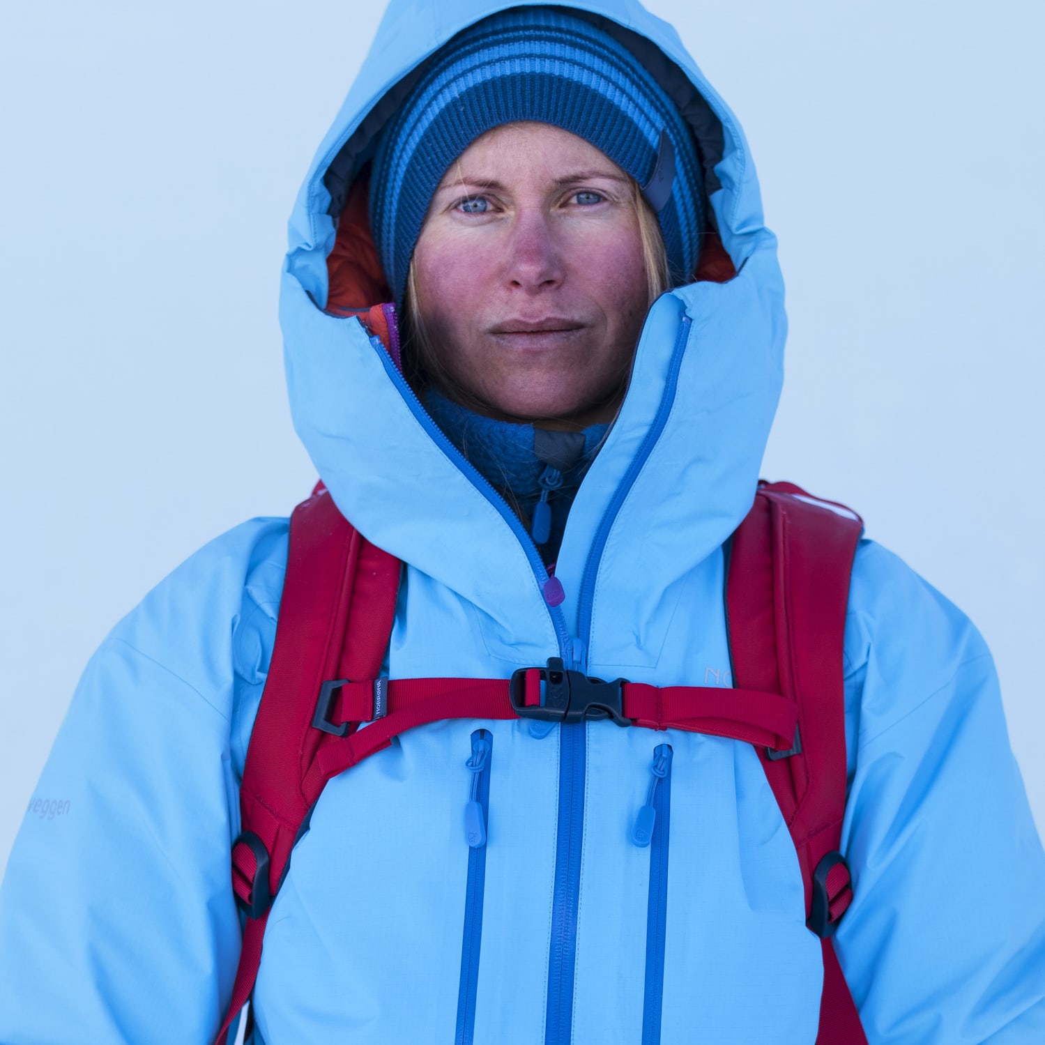 Portrait of a woman with blue eyes, wearing a blue Norrøna jacket, a blue striped hat and a red backpack, set against a snowy white background.