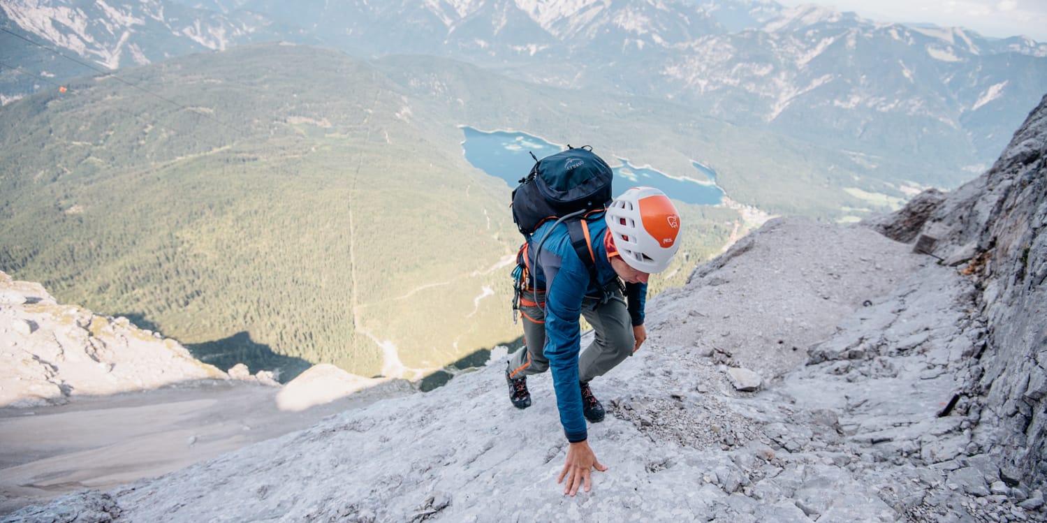 A climber in a Norrøna jacket and helmet ascends a rocky mountain face. A valley with a lake and mountain range is in the background.
