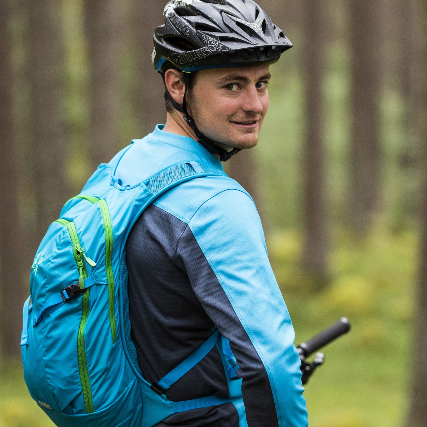 A mountain biker wearing a blue Norrøna shirt and backpack looks back in a forest.