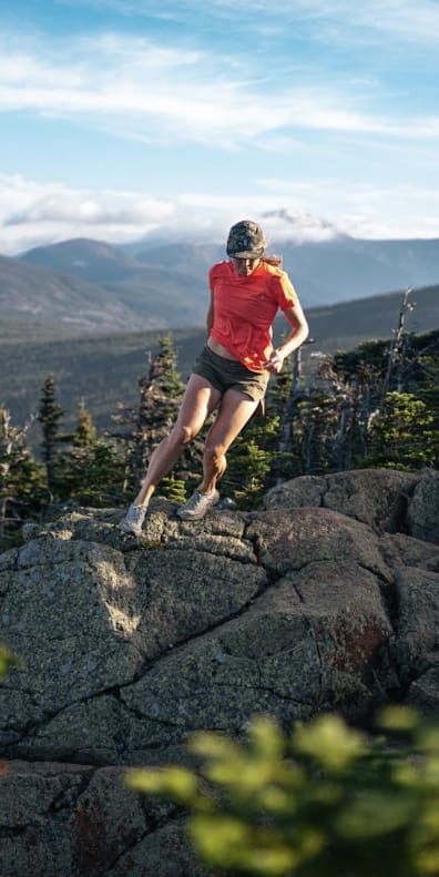 A woman in a Norrøna shirt runs across rocky terrain with a mountain range in the distance.