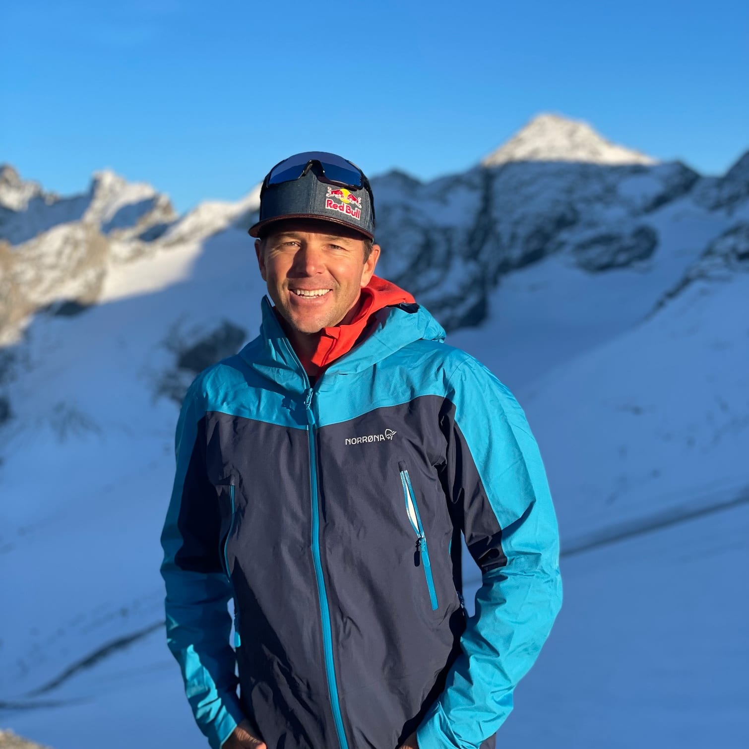 A smiling man with a Red Bull cap stands in front of snow-covered mountains wearing a blue and black Norrøna jacket.
