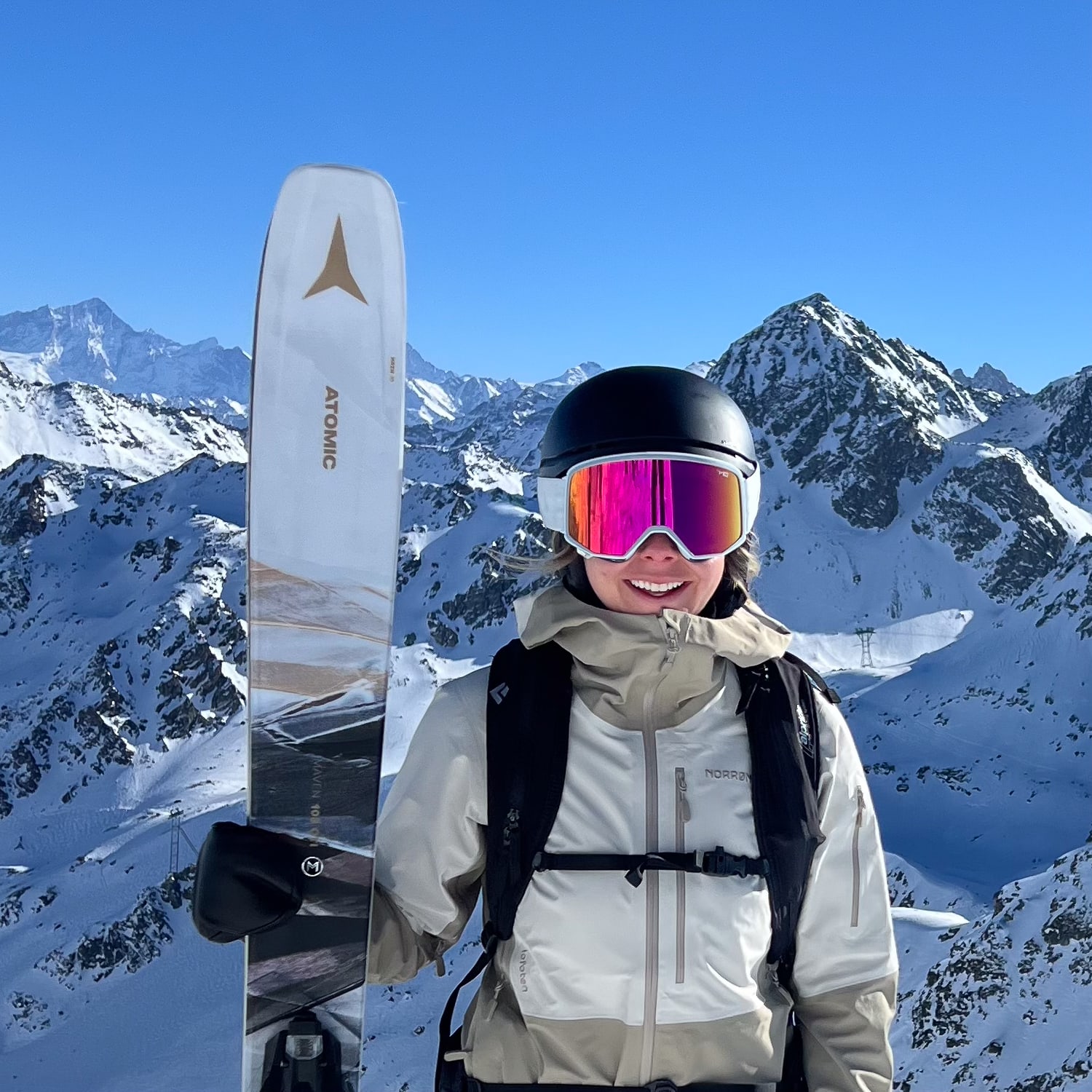 A person wearing a Norrøna jacket and pink ski goggles holds an Atomic ski against a snowy mountain backdrop. 
