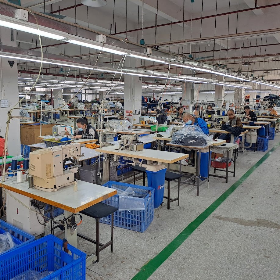 A factory floor with several rows of sewing machines. Workers are seated at the machines, sewing Norrøna clothing. There are blue plastic bins and black stools throughout the space. The floor has a green stripe running along the side of the image.
