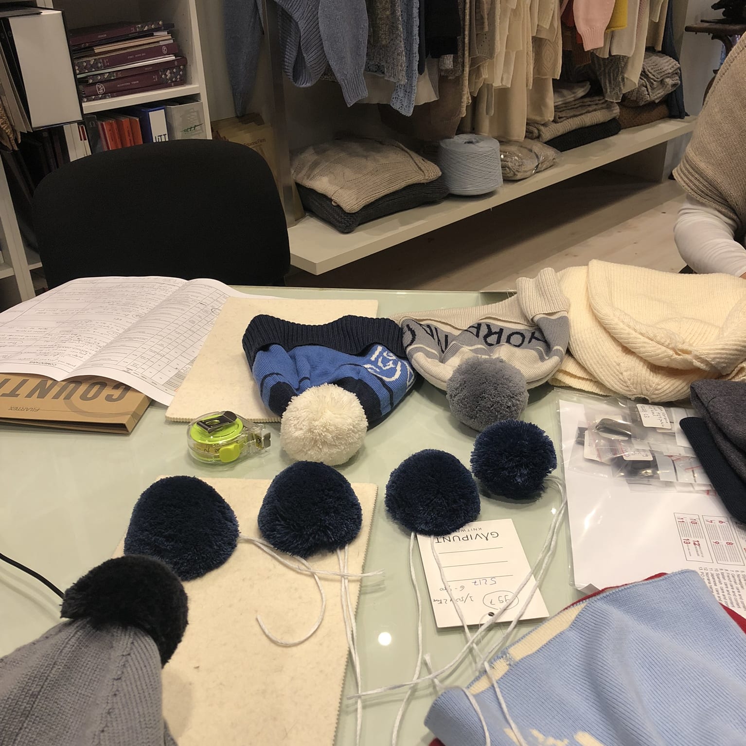 A table with various knitted hats, pom-poms, and other materials, including a tape measure and a tag that reads "GAVIPUNT KNITWEAR 52176-500".