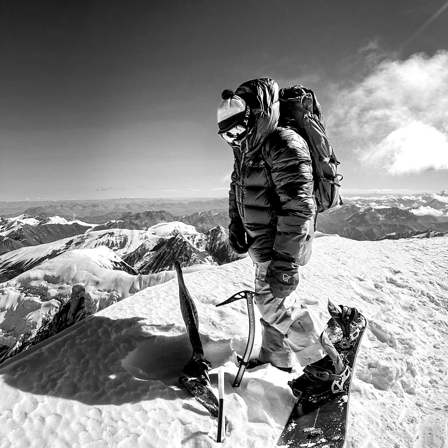 A person wearing a Norrøna jacket and pants stands on a snowy mountain top with a snowboard, ice axe, and backpack.