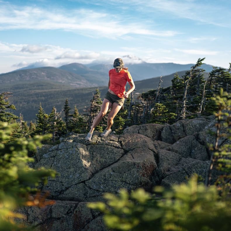 A woman in a Norrøna shirt runs across rocky terrain with a mountain range in the distance.