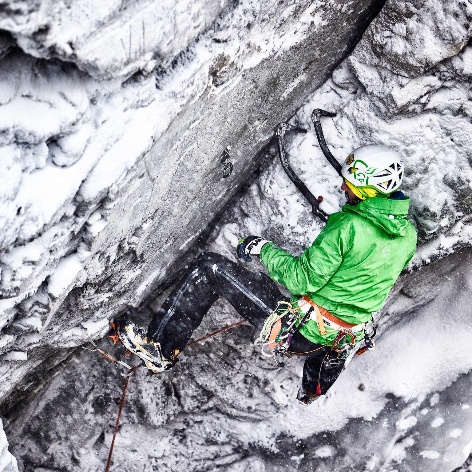 A climber in a green Norrøna jacket, helmet, and climbing gear, is ascending a snowy rock face using ice axes.