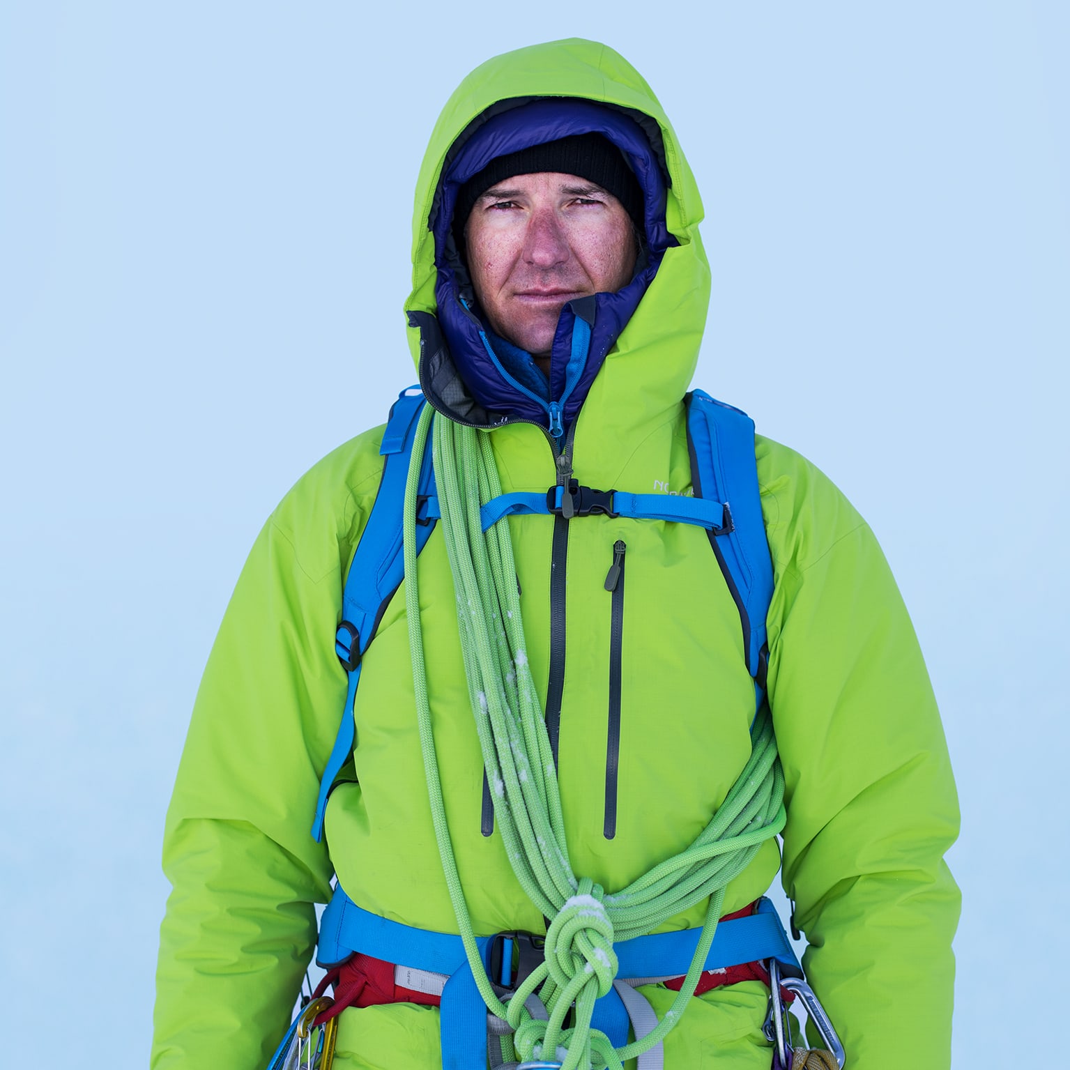 A climber with a rope on his shoulder wears a Norrøna hooded jacket, backpack, and climbing harness against a light blue background.