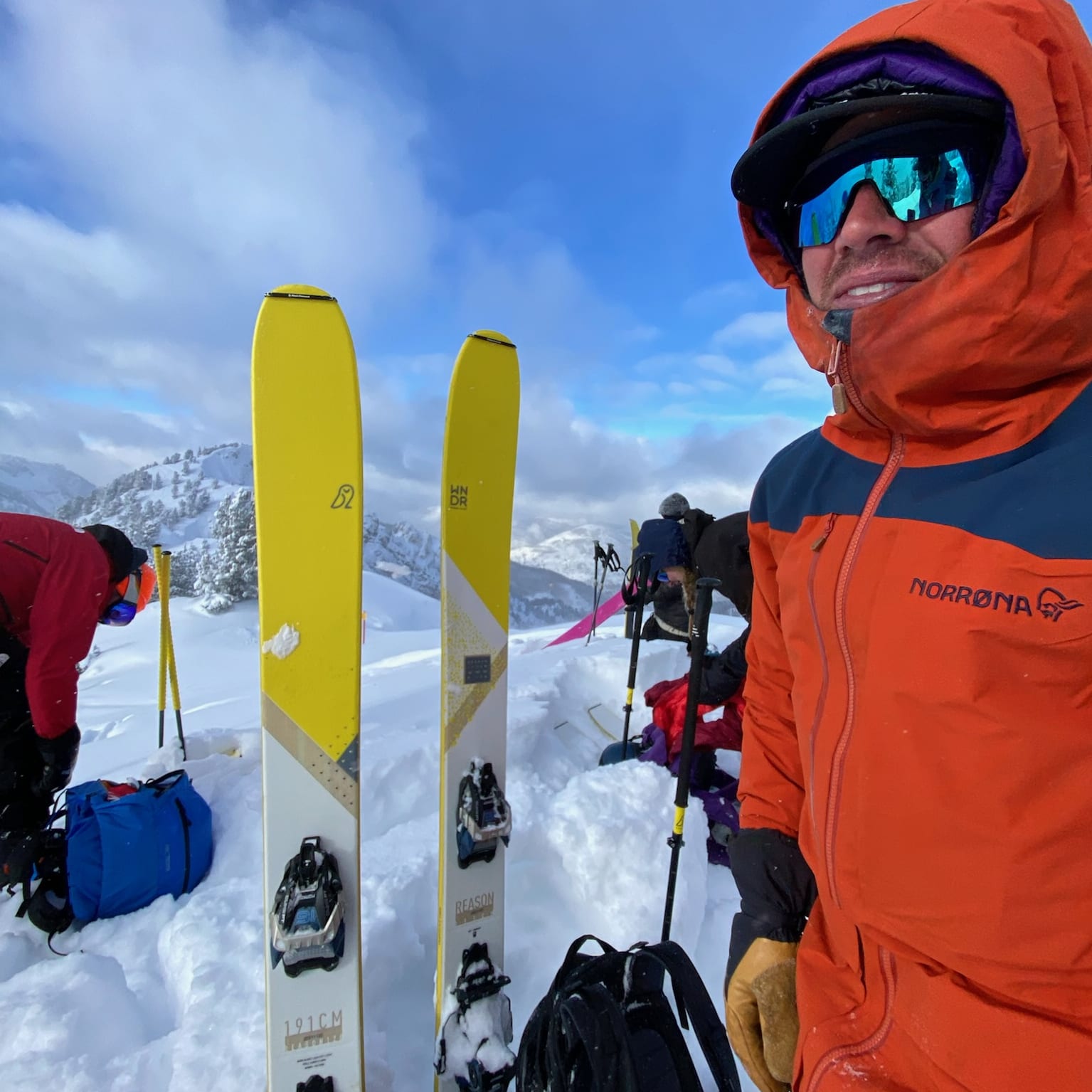 A skier with reflective goggles and a Norrøna orange jacket stands in snowy mountains with skis. Blue sky with clouds is in the background.
