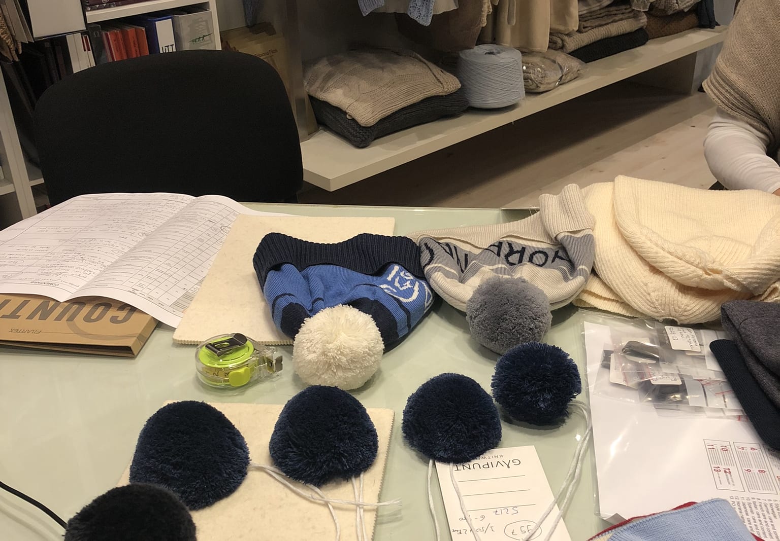 A table with various knitted hats, pom-poms, and other materials, including a tape measure and a tag that reads "GAVIPUNT KNITWEAR 52176-500".