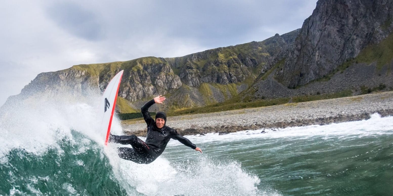 A surfer wearing a black Norrøna wetsuit rides a wave in front of a rocky coastline and mountains.