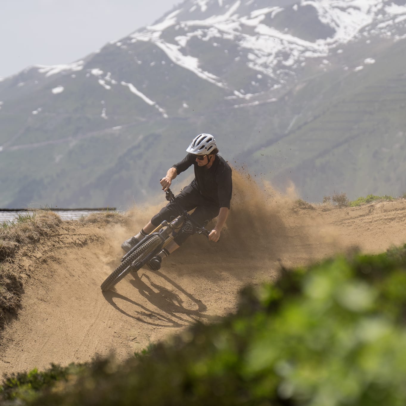 A person wearing a helmet rides a mountain bike down a dirt trail. The bike kicks up dust as it turns on a bend. The mountain bike rider is wearing Norrøna clothing.