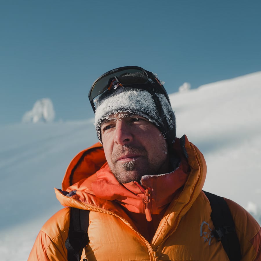 A man in a Norrøna jacket and hat stands in a snowy landscape.  The snow covers his hat and the top of his face. 
