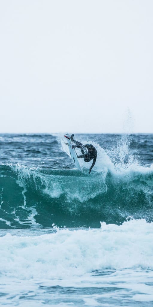 A surfer in a Norrøna wetsuit performs an aerial maneuver on a large wave. 
