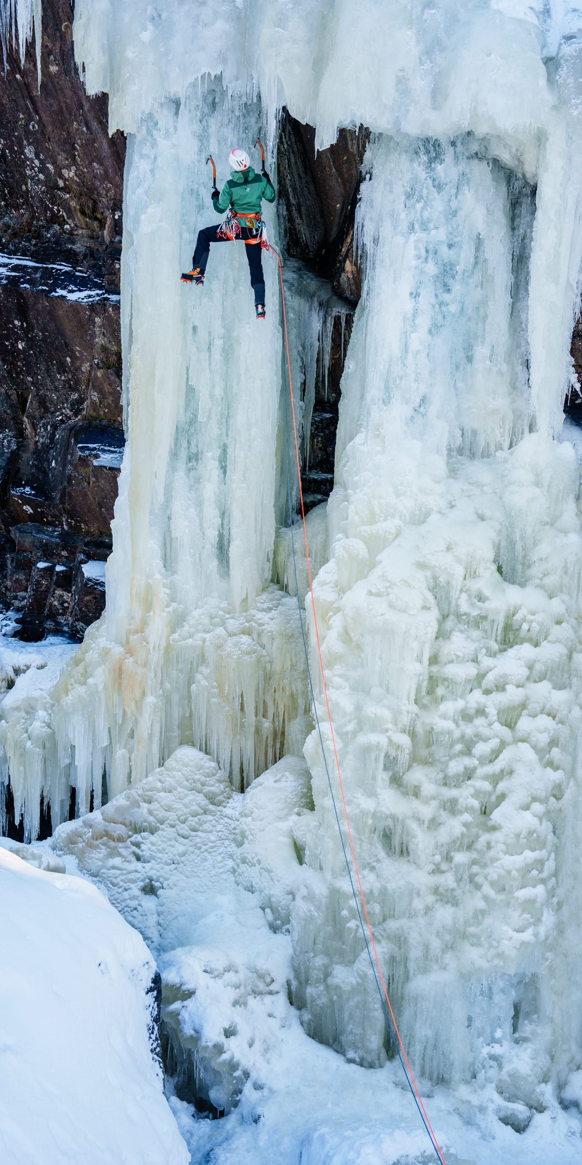 A climber ascends a frozen waterfall with ice axes and a Norrøna jacket. 

