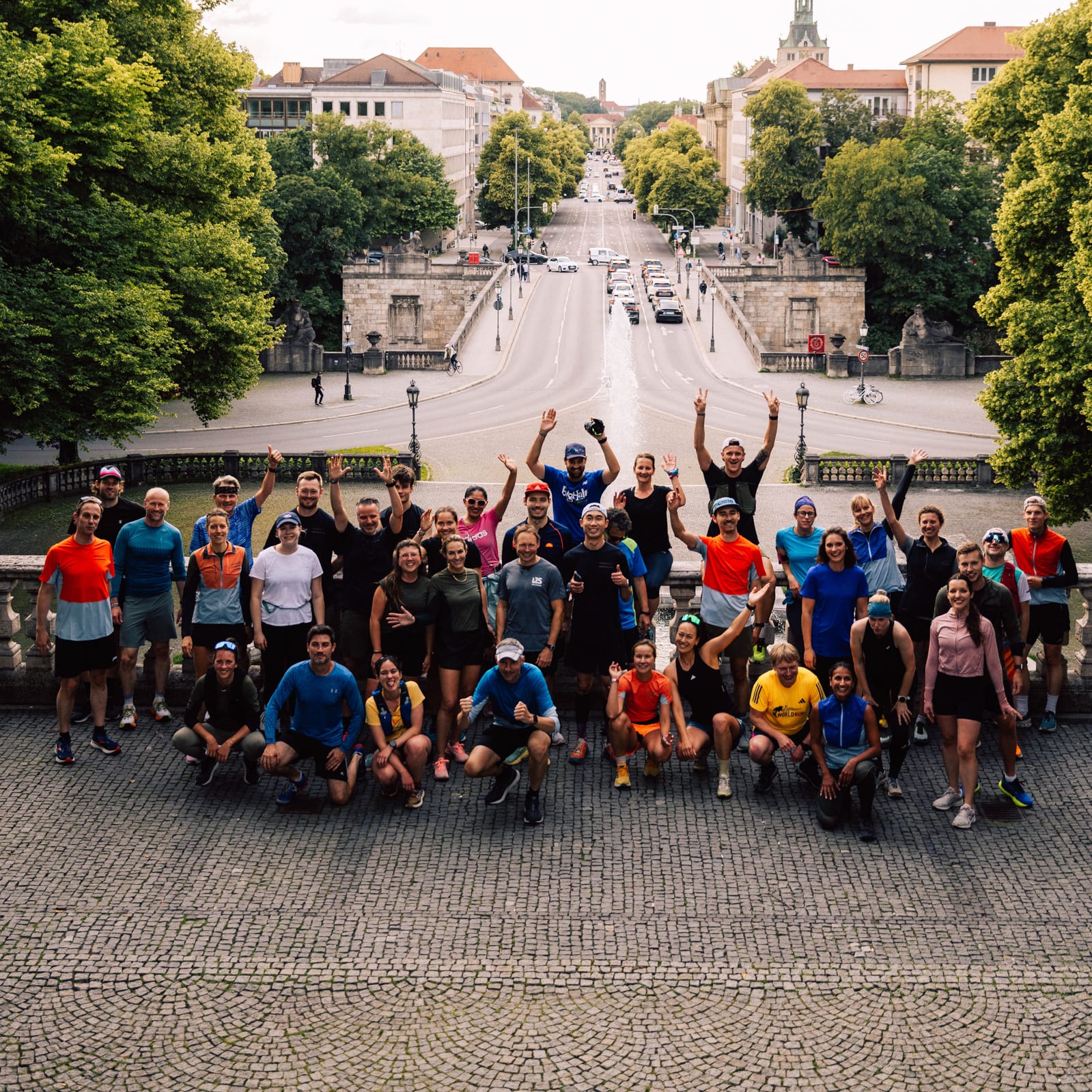 A group of people stand on a cobblestone path. Some are wearing Norrøna gear.  They are smiling and looking at the camera. The path leads to a street lined with trees and buildings.