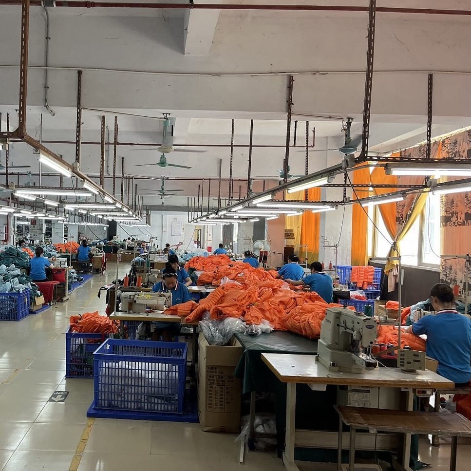 A large factory room with many people working at sewing machines. They are all wearing blue shirts and working on orange fabric. There are many blue plastic crates filled with fabric around the room.