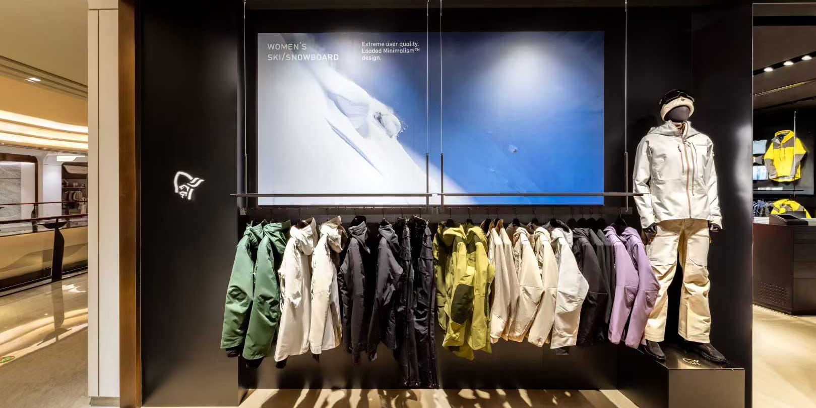 Store display showcasing Norrøna ski/snowboard jackets in various colors hanging below a mountain scene graphic. A mannequin wears a matching Norrøna jacket and pants.
