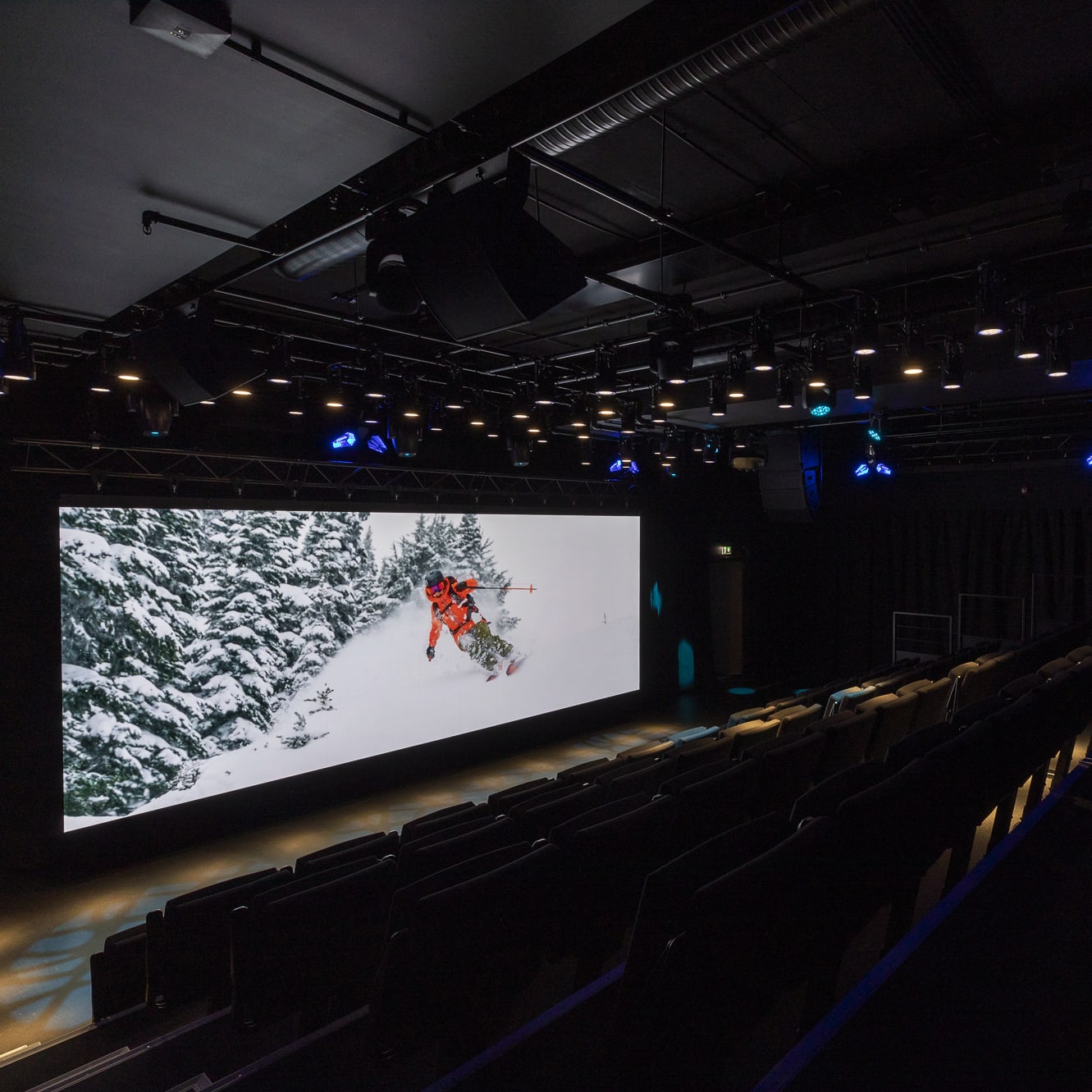 A large screen in a darkened auditorium showing a skier in a Norrøna jacket wearing a bright orange jacket skiing down a snowy mountainside. The auditorium has a large screen and many rows of seats.