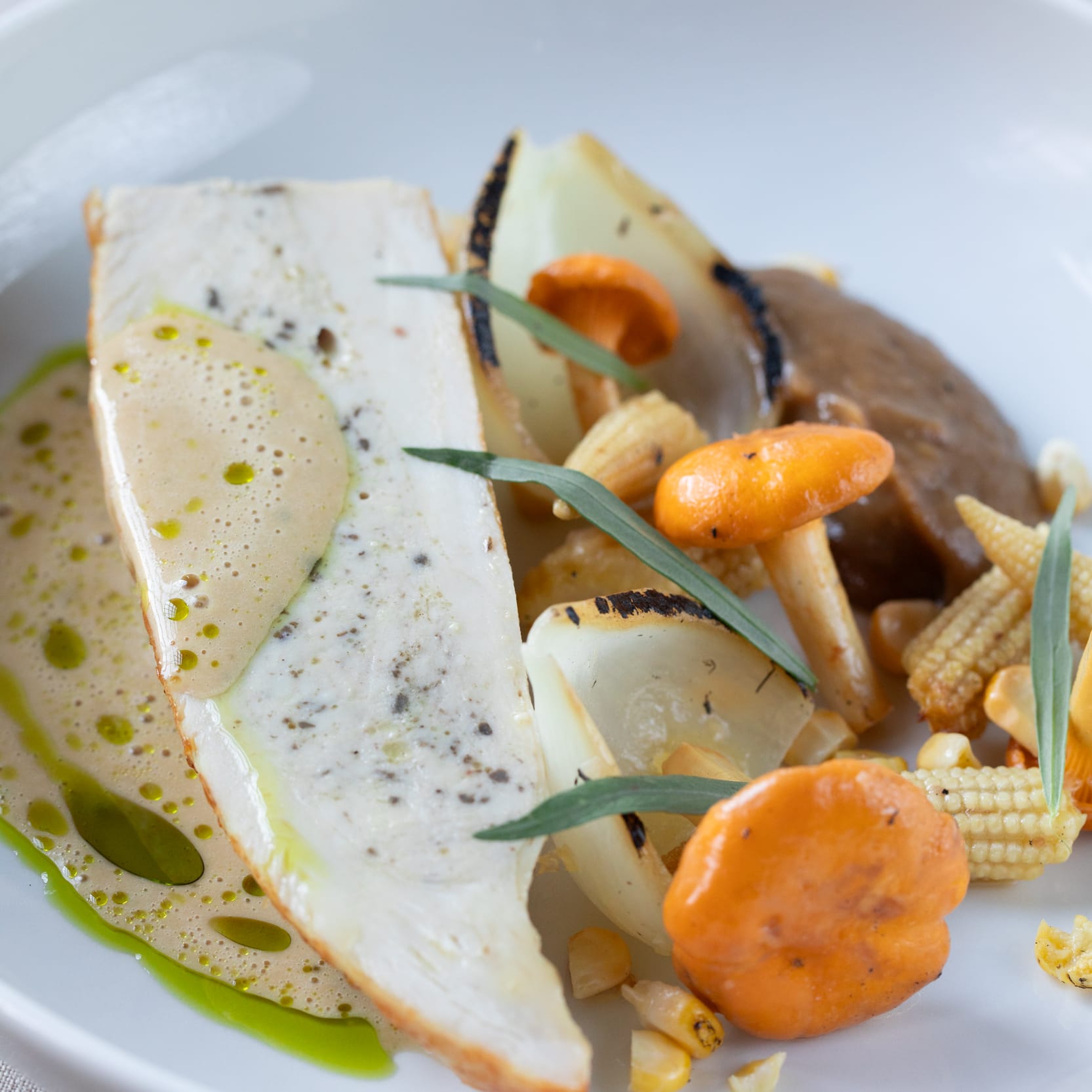 Close-up of a plate with a white fish fillet, chanterelle mushrooms, grilled onion, baby corn, and decorative sauces.