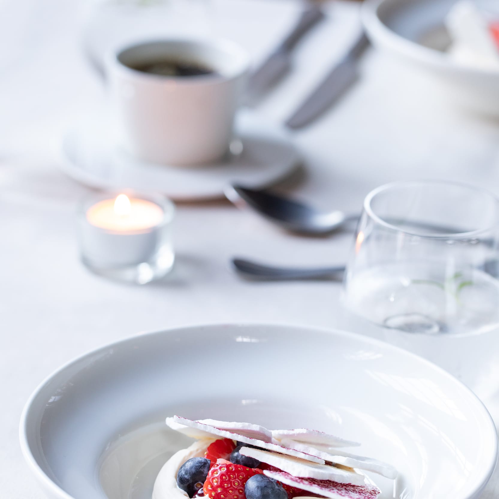 A dessert with berries and meringue on a white table, a cup of coffee, and a candle in the background.