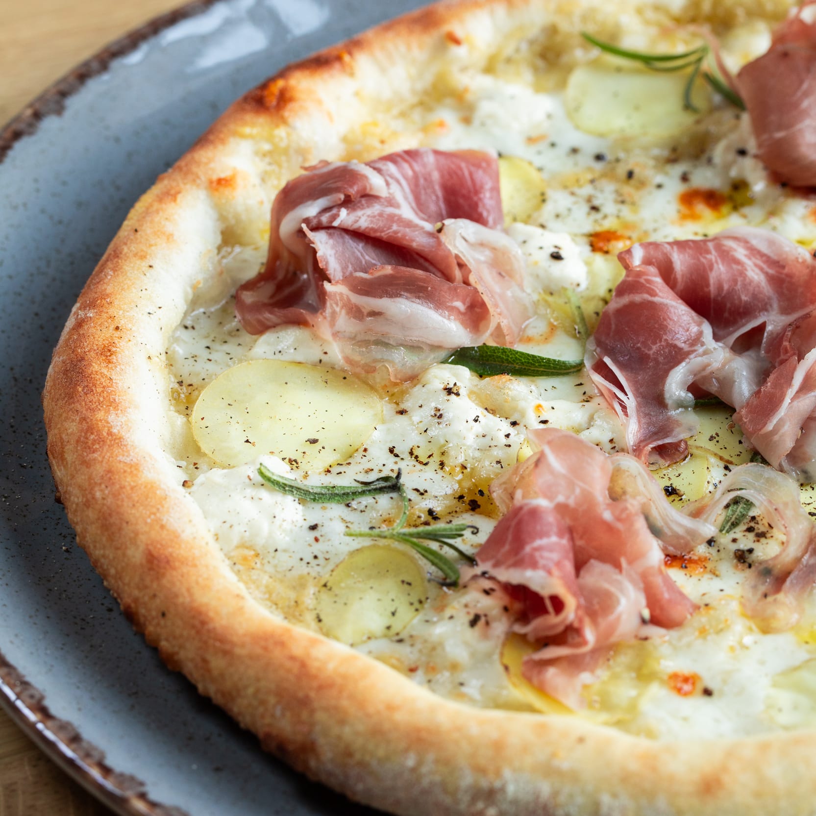 Close-up of a pizza on a gray plate, topped with cheese, potatoes, rosemary, black pepper and cured ham, resting on a wooden table.