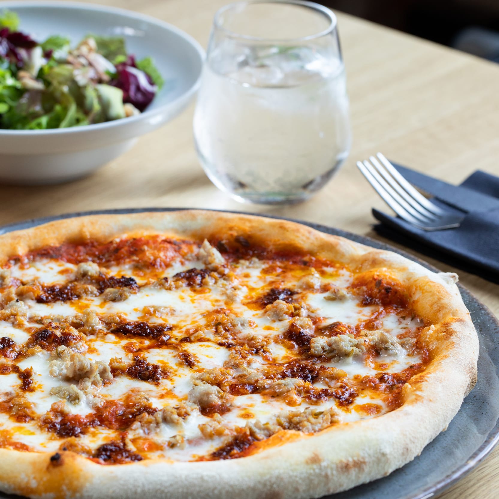 Pizza with sausage and tomato sauce, a salad, and a glass of water sit on a table.