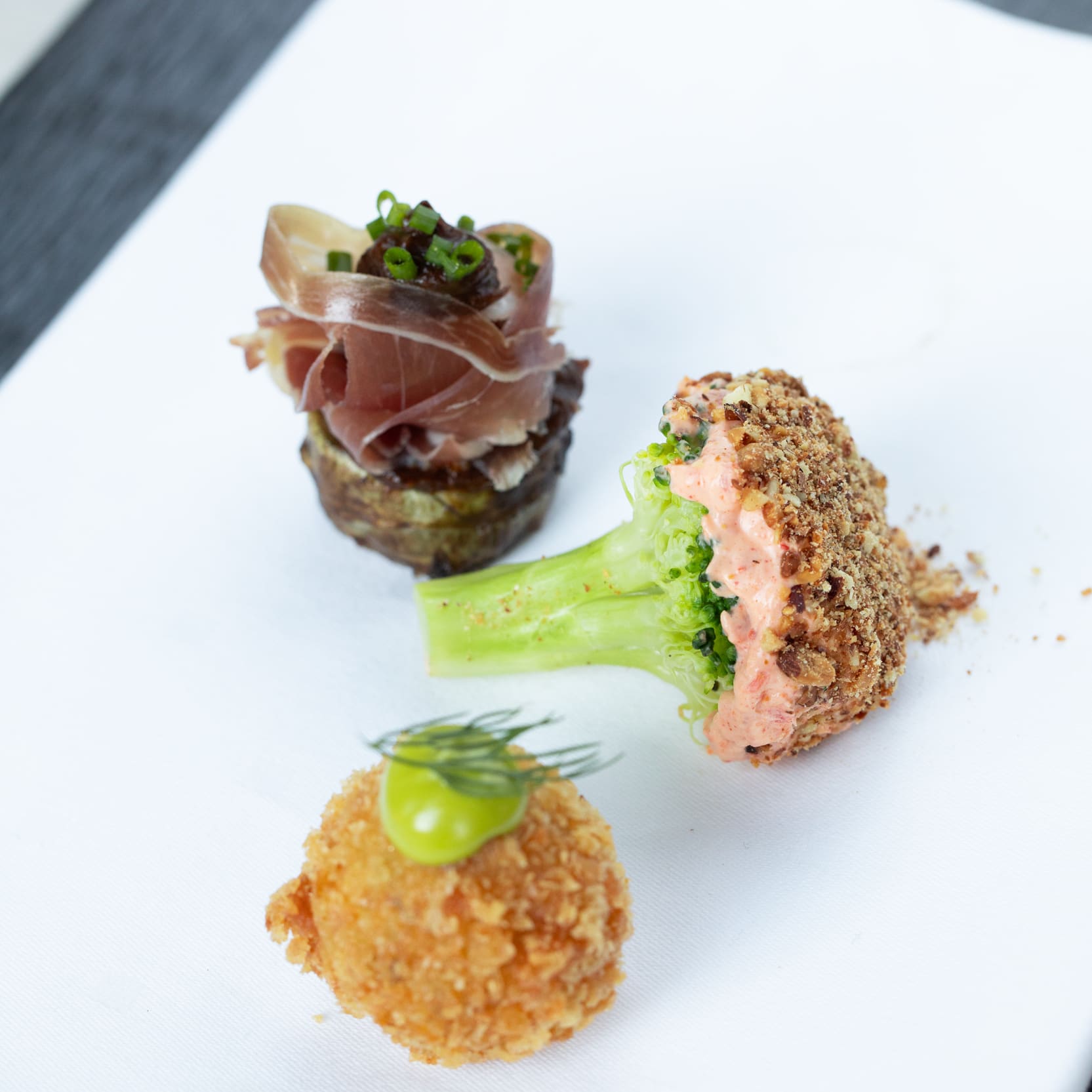 Three appetizers on a white cloth: prosciutto on potato, breaded broccoli with dip, and a fried ball with green sauce.
