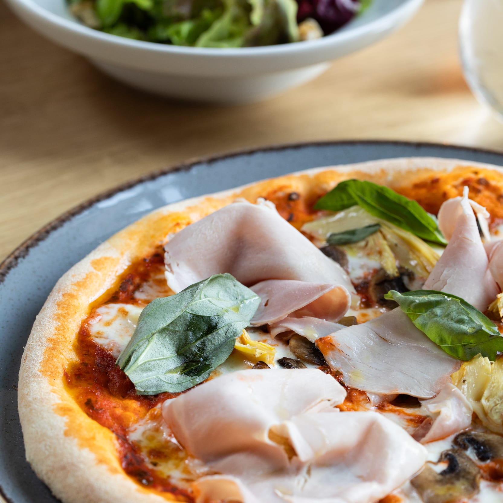 Overhead shot of a pizza on a gray plate, topped with ham, mushrooms, artichoke hearts, and basil. A salad is visible in the background.