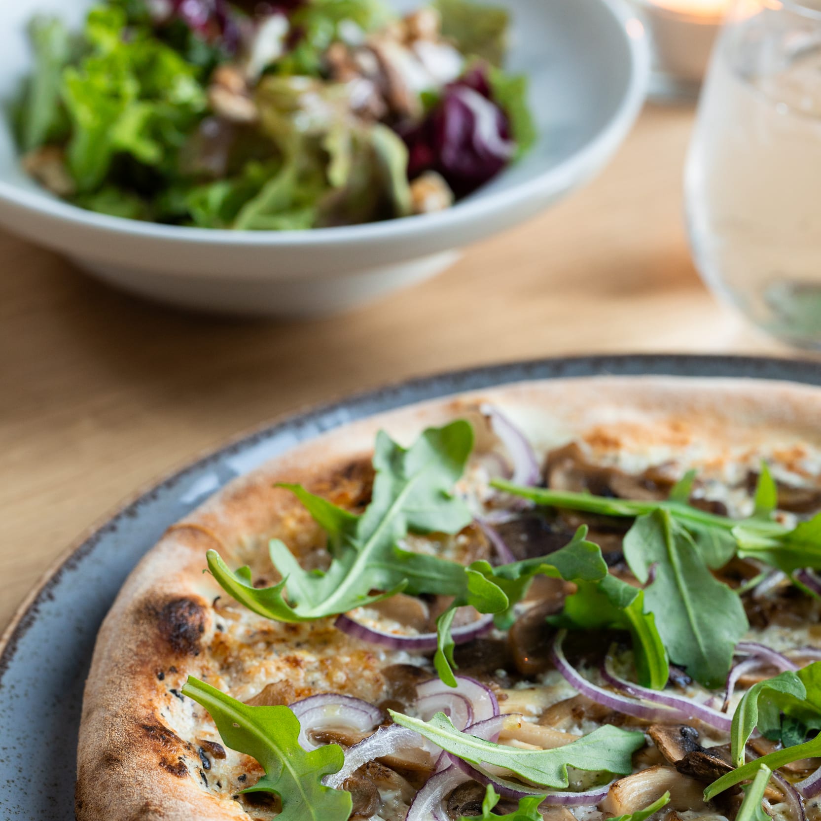 A mushroom and red onion pizza on a grey plate, with a salad and drink in the background.